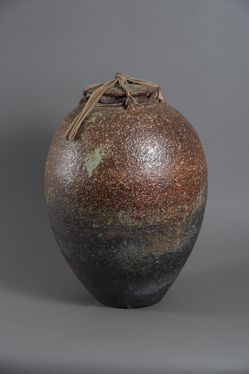 A large ceramic vessel with a brown "sugary" textured glaze. The vessel has a lid secured into place with leather chord and tassel. 