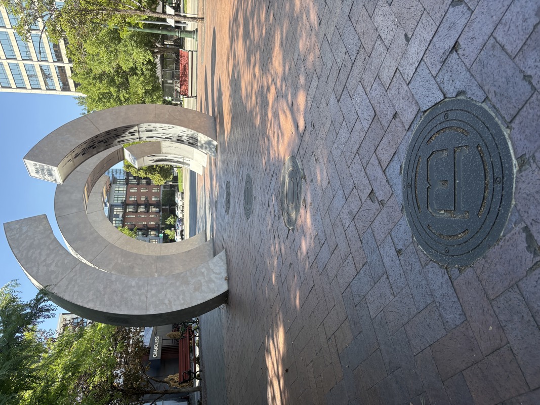 A sculpture made from brushed galvanized steel in the shape of three large semi-circular rings standing in a line parallel to one another. Lining the inner edges of the rings are inlays of clear acrylic plastic with screen prints depicting images and text referencing the history of Boise's Grove Street and the Boise Canal. 