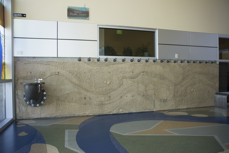 The interior side of the Boise WaterShed building. Along the bottom four feet of the building is a cement textural installation that resembles a birds-eye view or map of a river and its previous shorelines that have shifted over time. At the far left of the installation is Confluence, a drinking fountain made from a large metal watermain pipe. 
