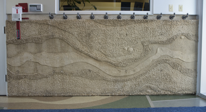 The interior side rear of the Boise WaterShed building. Along the bottom four feet of the building is a cement textural installation that resembles a birds-eye view or map of a river and its previous shorelines that have shifted over time. Along the top edge of the installation are eleven coat hangers made from metal water pipes.