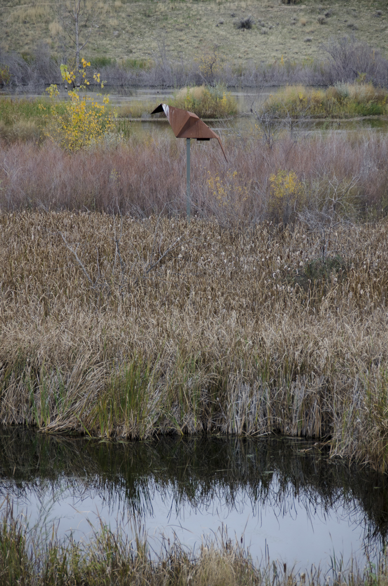 An abstracted geometric sculpture resembling a paper-folded origami bat in flight made of weathered steel, mounted onto a pole in the middle of a wetland.