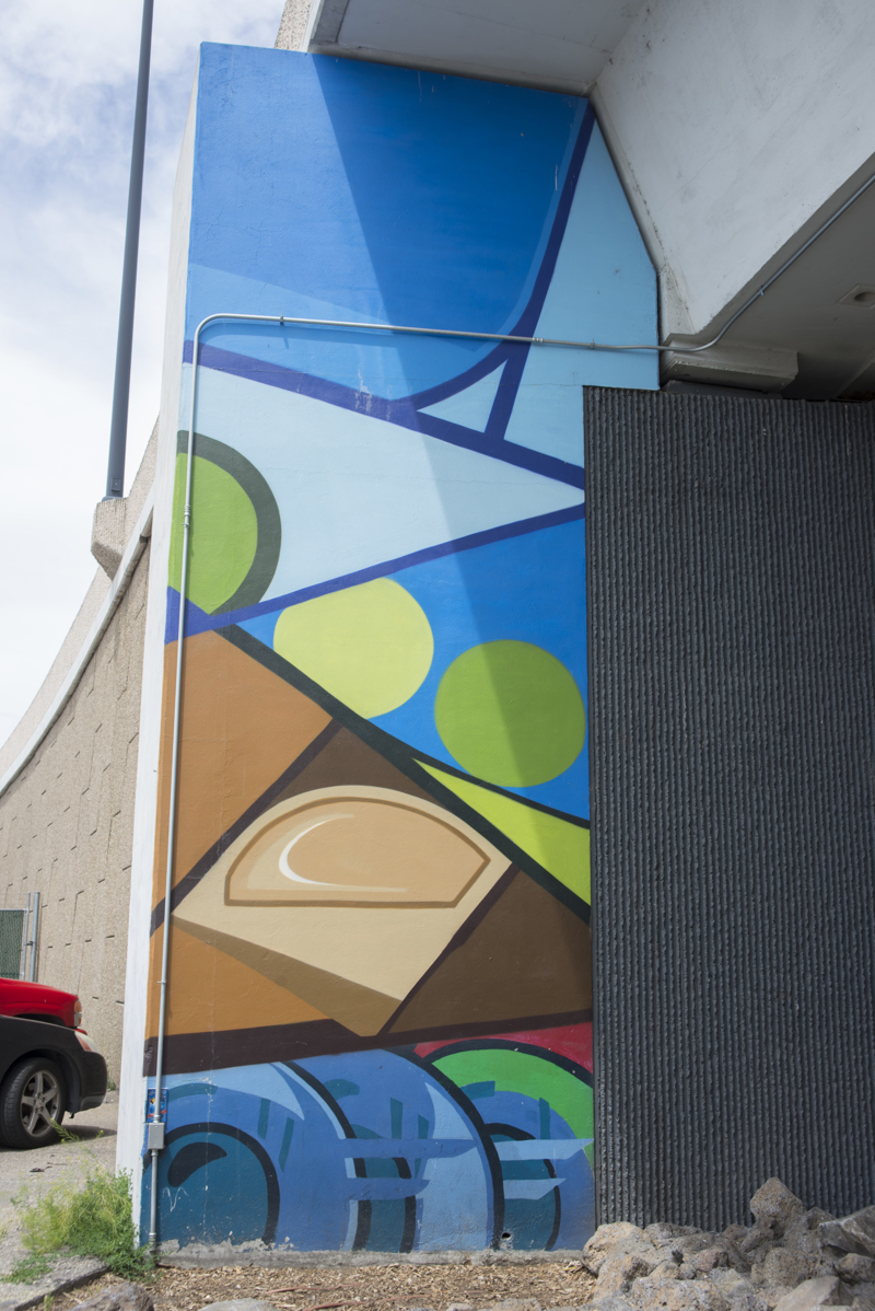 A mural painted in five parts on the wall of a highway overpass. All five sections are composed of geometric shapes with cartoon-like outlines and block colorations.