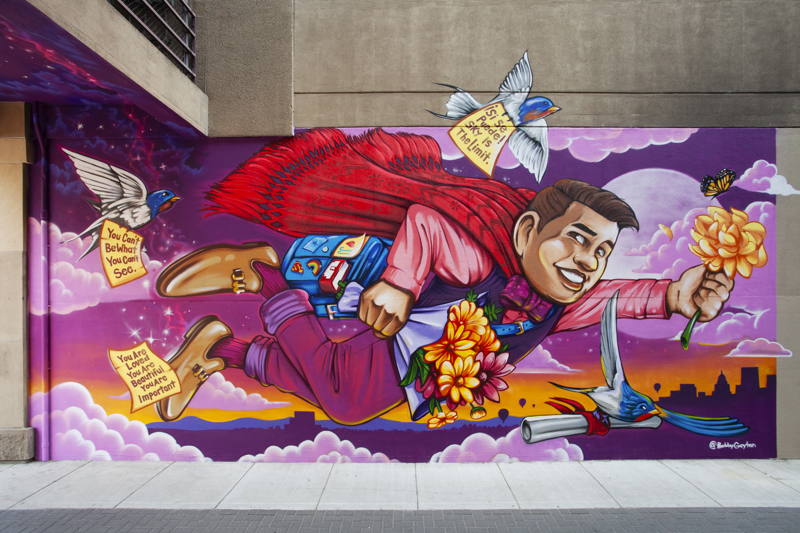 A painted mural that spans both sides of an alleyway. On one side is a dapperly dressed figure flying over the Boise skyline. They hold flowers and are surrounded by butterflies and swallows who carry notes of encouragement and affirmation. On the other side is an arrangement of flowers with butterflies, a hummingbird, a candle, and papel picado. On the far left is the word "Amore". 