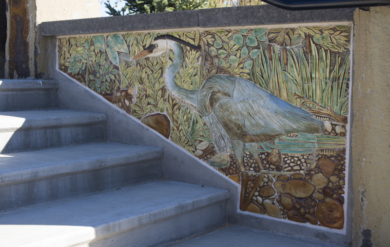 An inner wall of an exterior stairwell made with a sandstone top and a base decorated with painted low bas-relief ceramic sculptures depicting a great blue heron standing on the edge of a river.