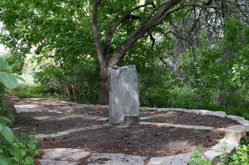 A short basalt stone column situated in a ring of flagstone. On top of the basalt column are four small patinated bronze-cast naturalistic sculptures attached to the top. The small sculptures are a feather, a nest of eggs, a charred branch, and a bone.