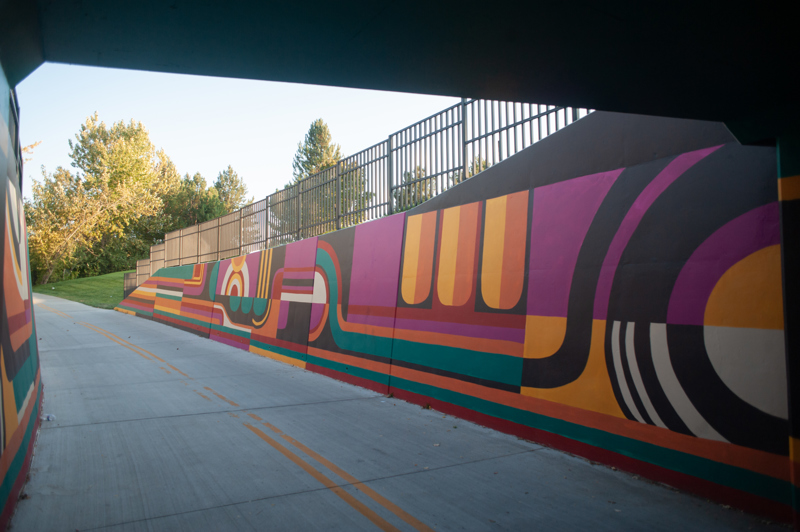 A mural on the retaining walls of a bike path heading into a tunnel. The design consists of lines and abstract shapes in the colors black, white, red, orange, yellow, teal, and magenta.