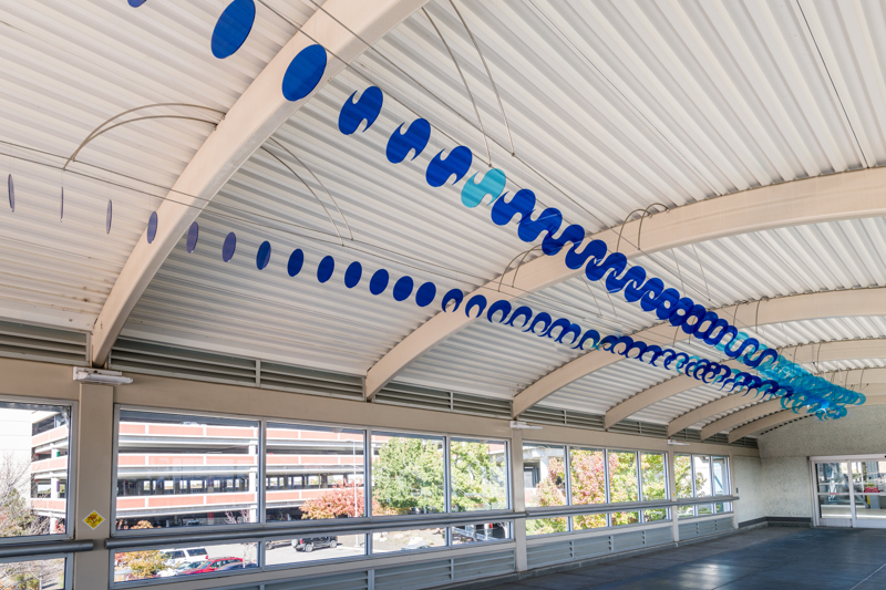 A series of 140 clear blue plastic acrylic cutouts that are in the shapes of exhaust whips that collectively resemble a long contrail spanning the length of a pedestrian sky bridge connecting the airport with a parking garage. 