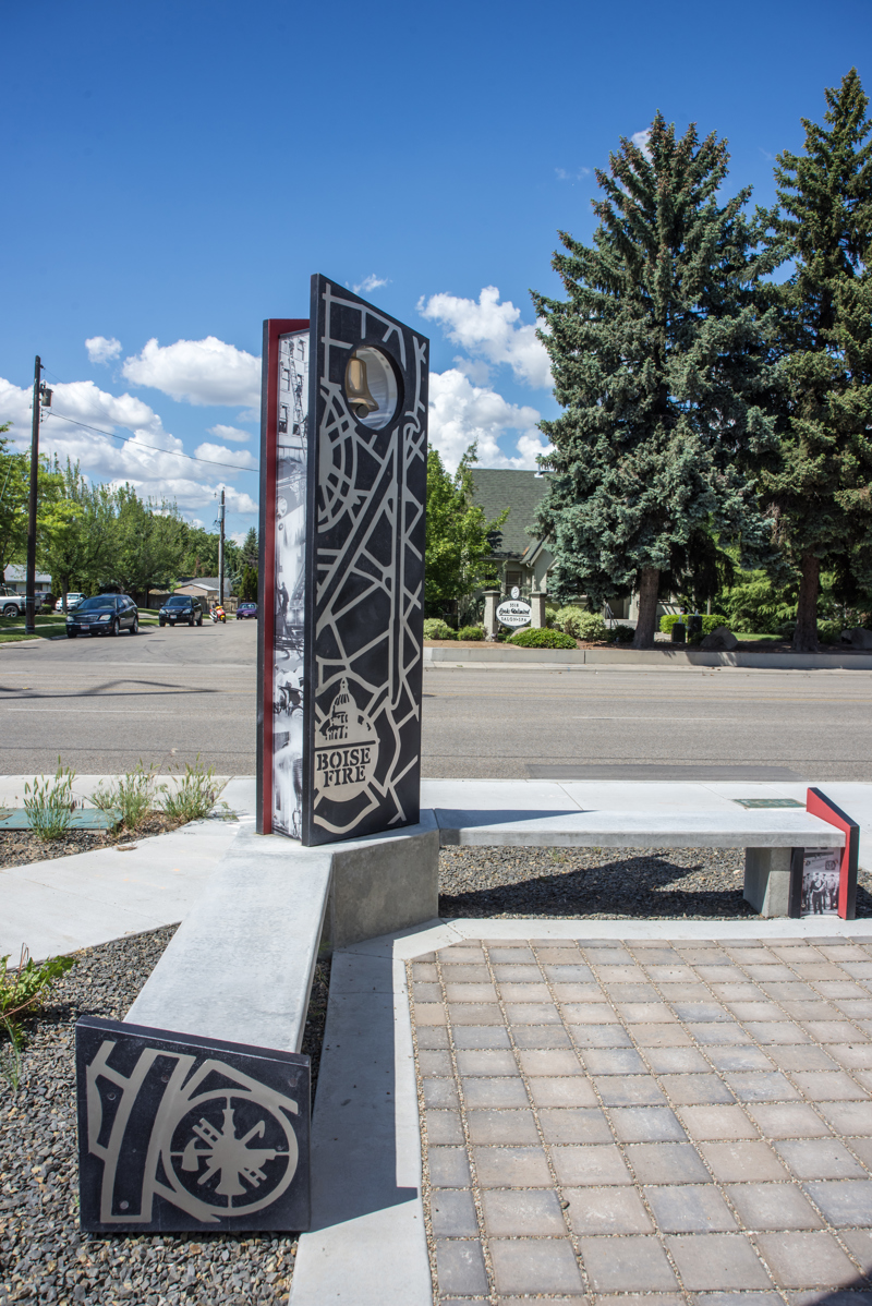 A corner bend made of cement, composite panels with printed images, and stainless steel cutouts of symbols and maps relating to Boise's first responders, namely firefighters. At the corner of the bend is a standing panel with a brass bell at its top. 