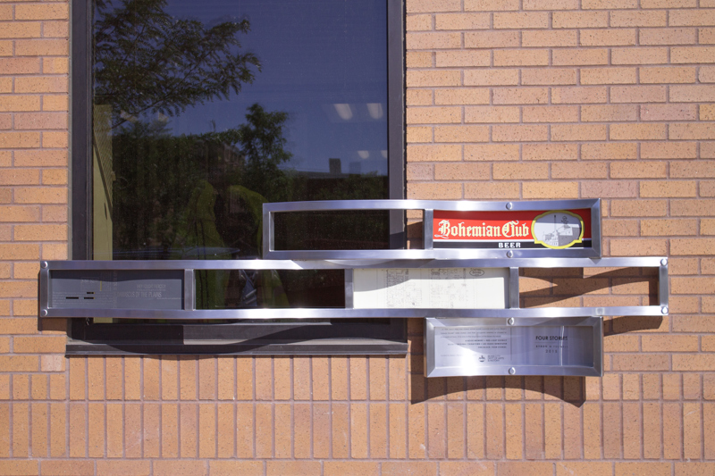 Six groupings of wavy stainless steel frames affixed to the exterior red brick wall of Boise City Hall. Some frames contain written and visual content with Chinese figures and historical references.