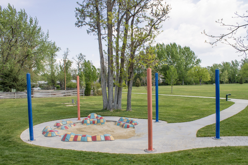 A small sand pit surrounded by sitting mounds covered by blue, yellow, and red ceramic tiles. The pit is encircled by brick and cement pathways leading off in different directions. Along the path are tall blue and red poles.