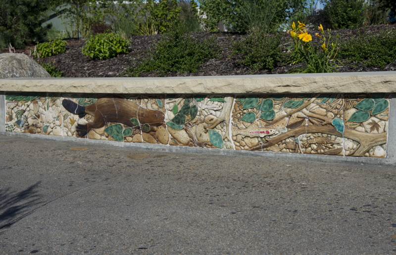 A flower bed retaining wall bench with a base decorated with painted low bas-relief ceramic sculptures depicting a beaver, fish, and crawfish swimming in a river.