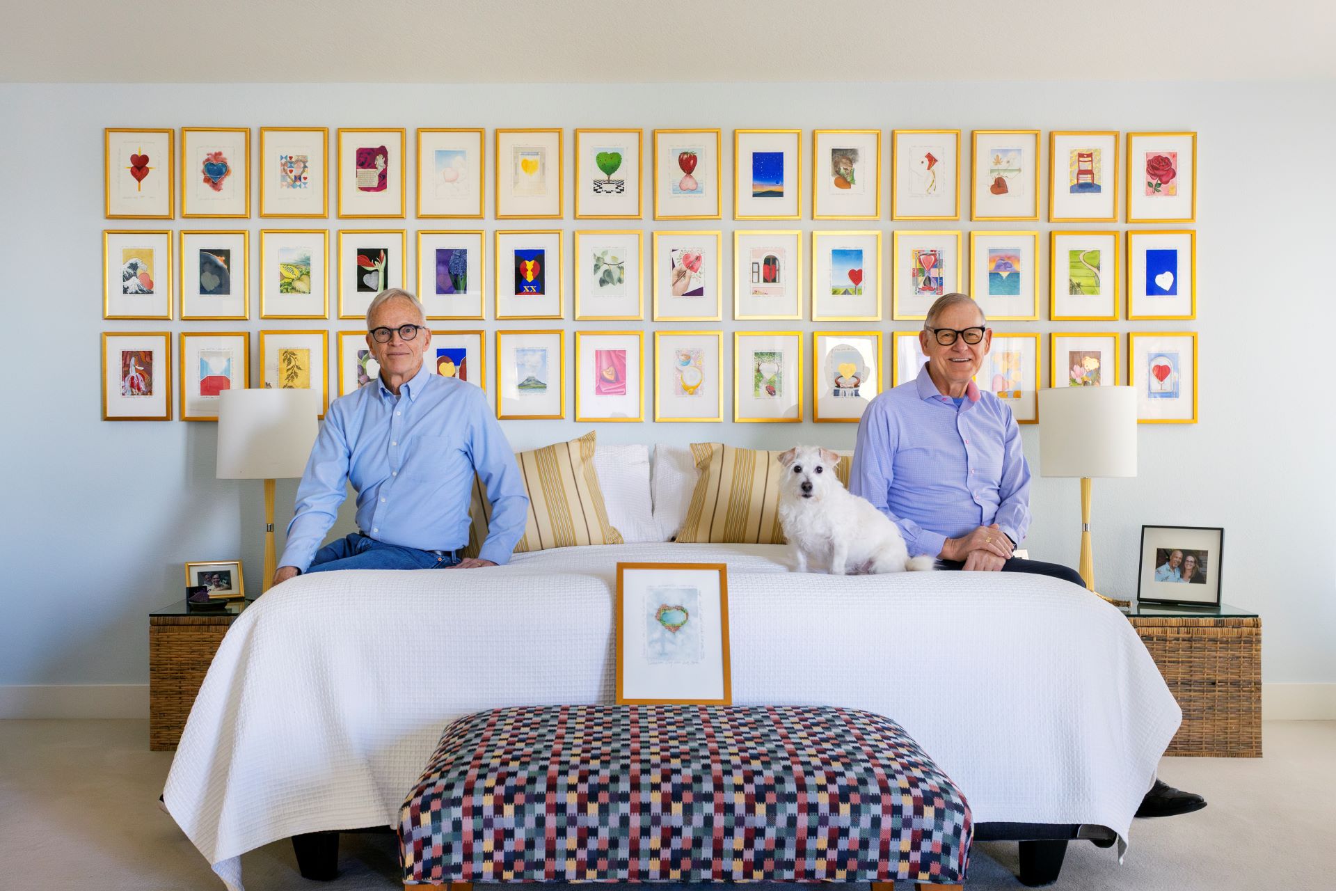 Two men sit on opposite edges of a bed, small dog in between, with many framed art works on the wall in the background.