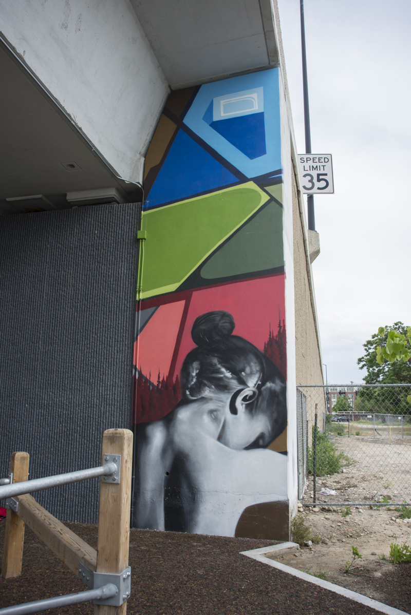 A mural painted in three parts on the wall of a highway overpass. On the left is the image of a yellow bird in flight, overlapping a leafy branch and a woman's bust. In the middle is an image of a woman in side profile with flowers in her hair. On the right is an image of a woman's upper back as she looks down. 