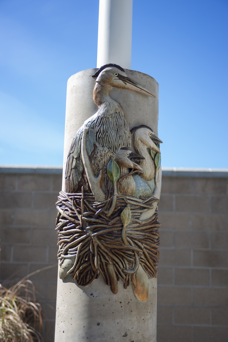 A cement column decorated with a ceramic mosaic relief sculpture of a nesting great blue heron and chicks.