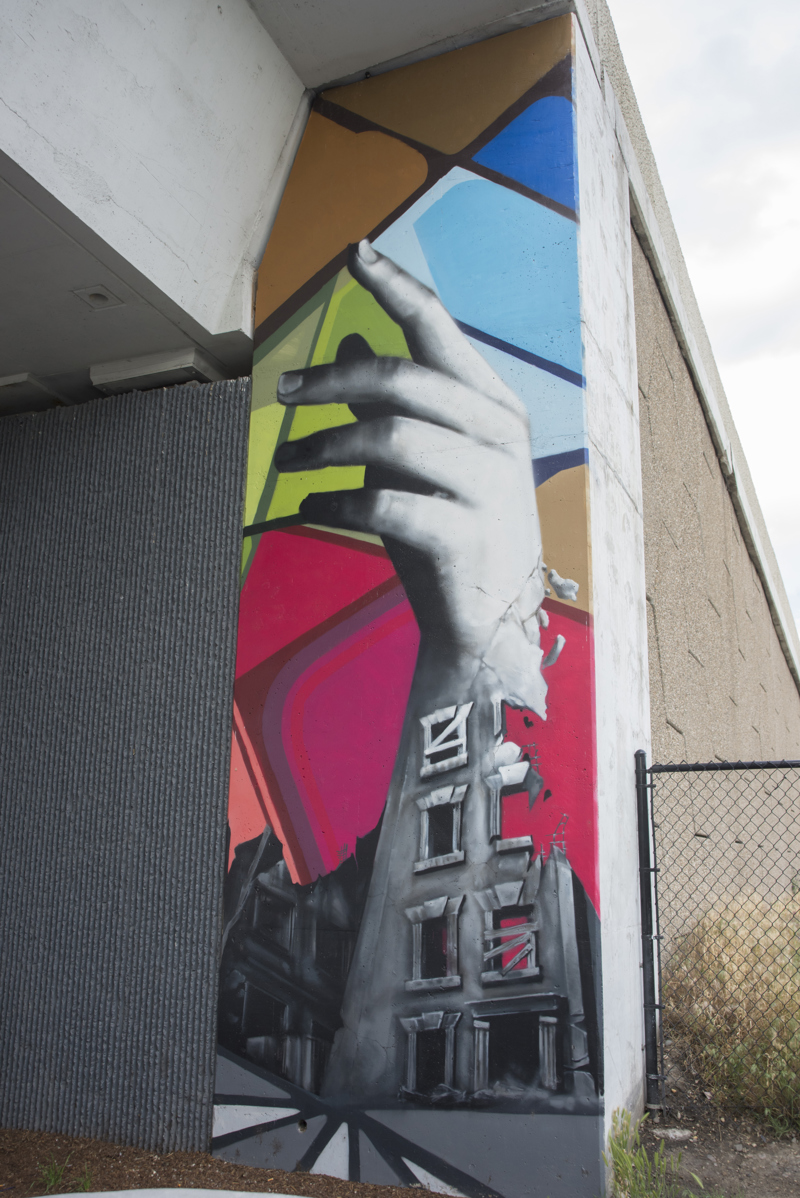 A mural painted in three parts on the wall of a highway overpass. On the left is the image of a pair of hands holding soil that slips through its fingers, turning into building rubble. In the middle is an image of a single eye situated beneath the arch of a bridge. On the right is an image of a hand and forearm turning into the crumbling facade of an abandoned building. 