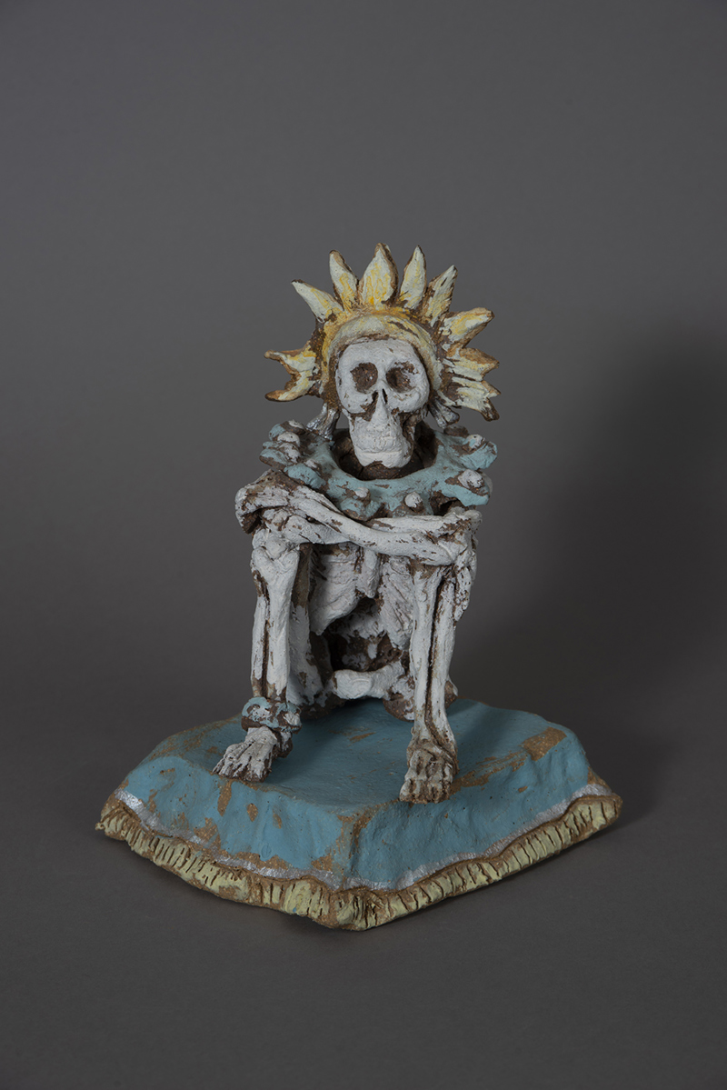 A sculpture of a skeleton with crossed arms, seated on a pillow with fringe. On the skeleton's head is a crown resembling a sunflower.