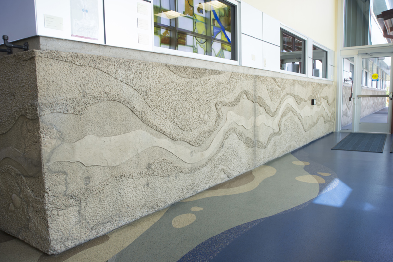The interior front entrance of the Boise WaterShed building. Along the bottom four feet of the building is a cement textural installation that resembles a birds-eye view or map of a river and its previous shorelines that have shifted over time. 