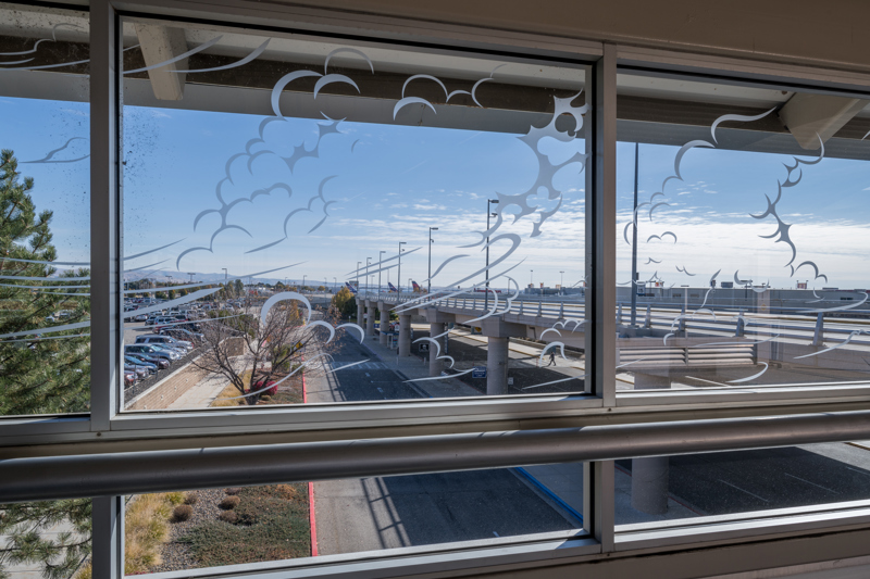 A series of windows located in the pedestrian sky bridge connecting the Boise Airport with a parking garage. On the windows are white vinyl stickers in the shapes of wisping clouds.