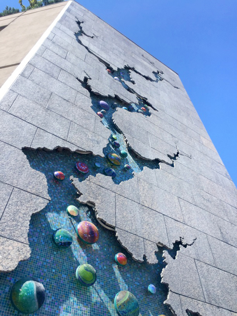 A large scale installation spanning nearly fifty feet of a building corner made up of rectangular granite slabs. Running up the center of the wall is a cutout shape of a large shape resembling a crack or a river and its tributaries. Recessed in this "crack" is a blue glass-tiled surface and circular, colorful blown glass components. 