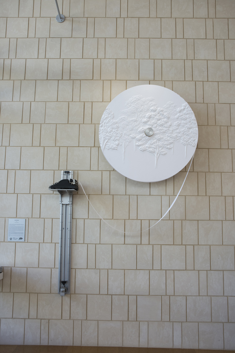 An antique typewriter set atop of a shelf that can be raised or lowered along a crankshaft attached to a sandstone wall. The typewriter is fed from a large roll of paper also attached to the wall. The surface of the paper roll features a low-bas relief carving of poplar trees. The typewriter has been lowered and a person in a blue dress is using it to type.