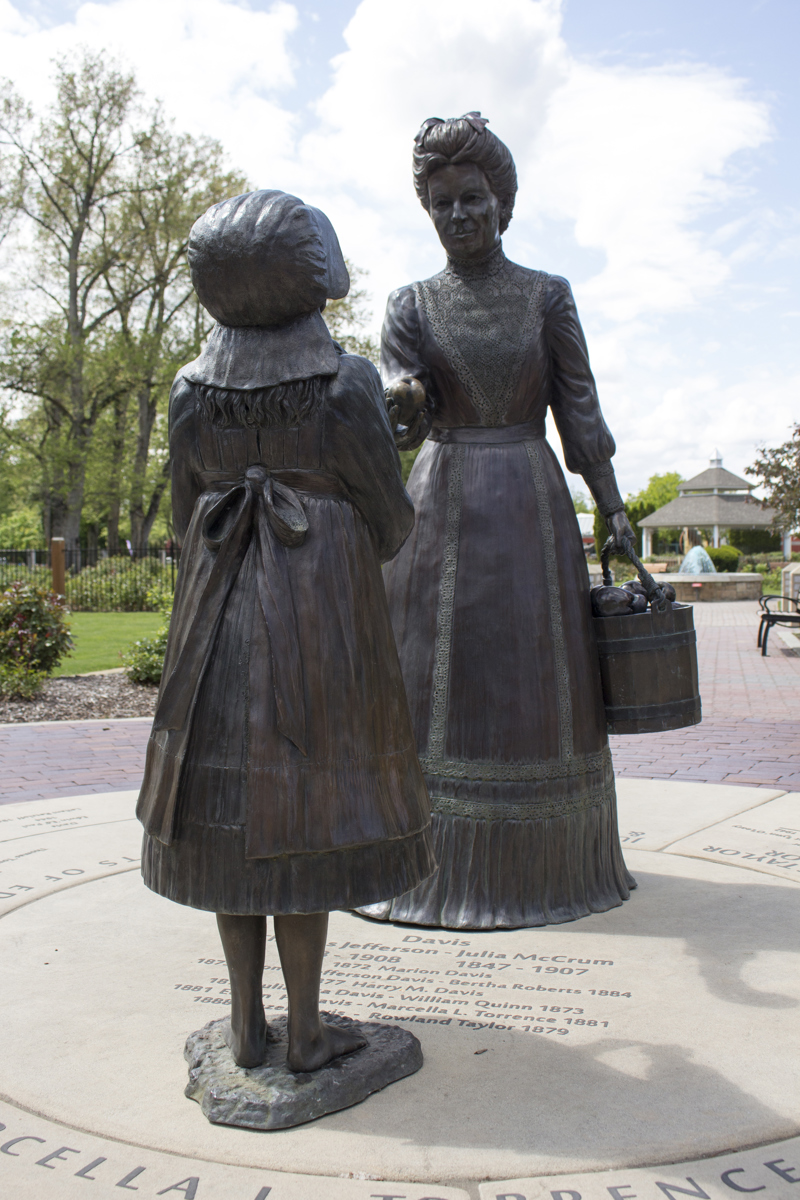 A life size bronze sculpture of a woman dressed in Victorian Era clothing (Julia Davis) holding a bucket of apples, and handing an apple to a young girl.