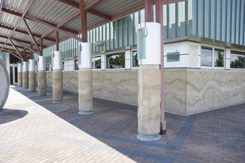 The exterior front of the Boise WaterShed building which has seven columns. Along the bottom four feet of the building and columns is a cement textural installation that resembles a birds-eye view or map of a river and its previous shorelines that have shifted over time.