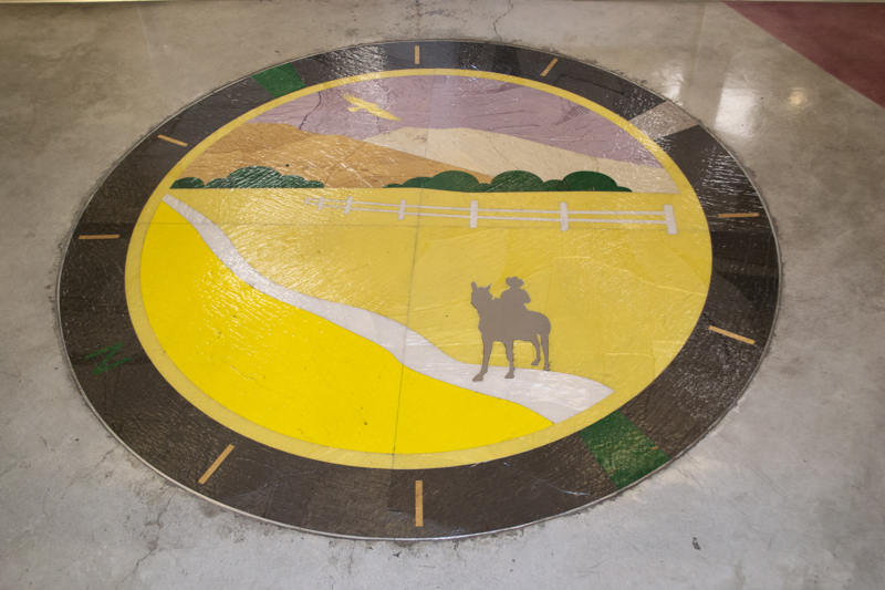 A floor inlay medallion depicting a person riding a horse along a path in a field. Around the outside of the medallion are the cardinal directions with the letter N indicating north. 