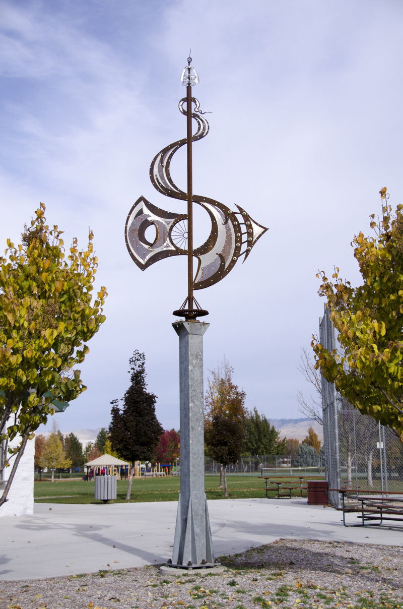 A tall metal sculpture resembling a weather vane. The base column is made from galvanized steel. The sculptural element at the top is made from a mixture of metals including Corten steel, stainless steel, and aluminum. It resembles a decorative letter S.