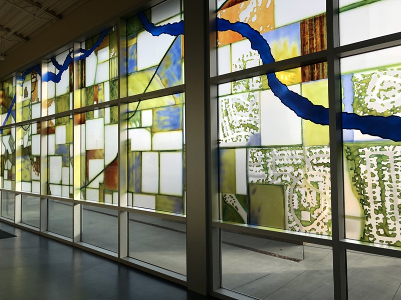 A window installation resembling stained glass depicting a birds-eye view of a blue river flowing through the geometric rectangular shapes, some that looks like fields and housing communities.