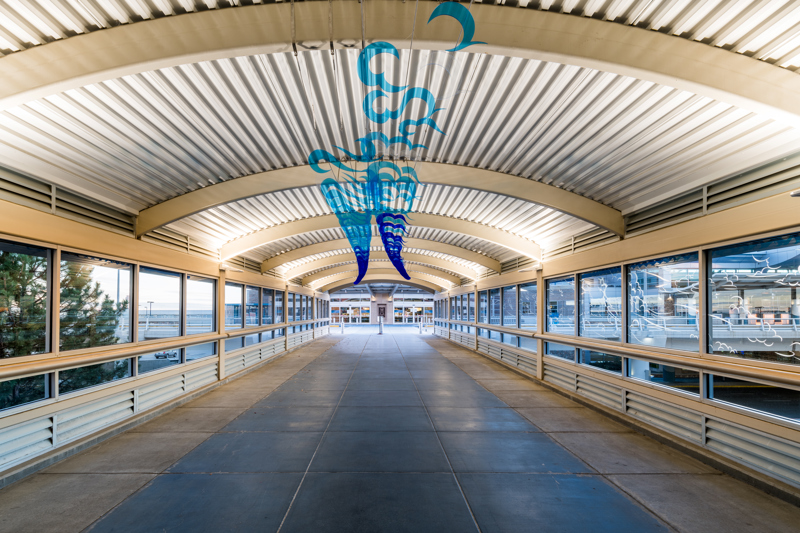 A series of 140 clear blue plastic acrylic cutouts that are in the shapes of exhaust whips that collectively resemble a long contrail spanning the length of a pedestrian sky bridge connecting the airport with a parking garage. 