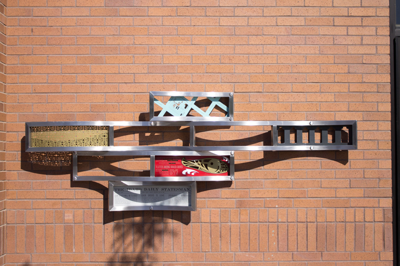 Six groupings of wavy stainless steel frames affixed to the exterior red brick wall of Boise City Hall. Some frames contain written and visual content with Chinese figures and historical references.