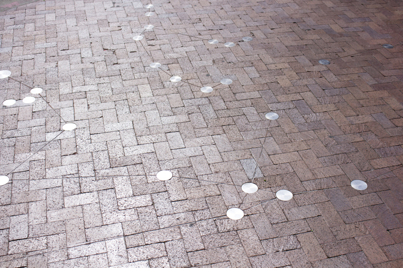 A collection of silver medallions inlaid into a cobble brick walkway. Lines are cut shallowly into the brick, connecting the medallions to form constellations.