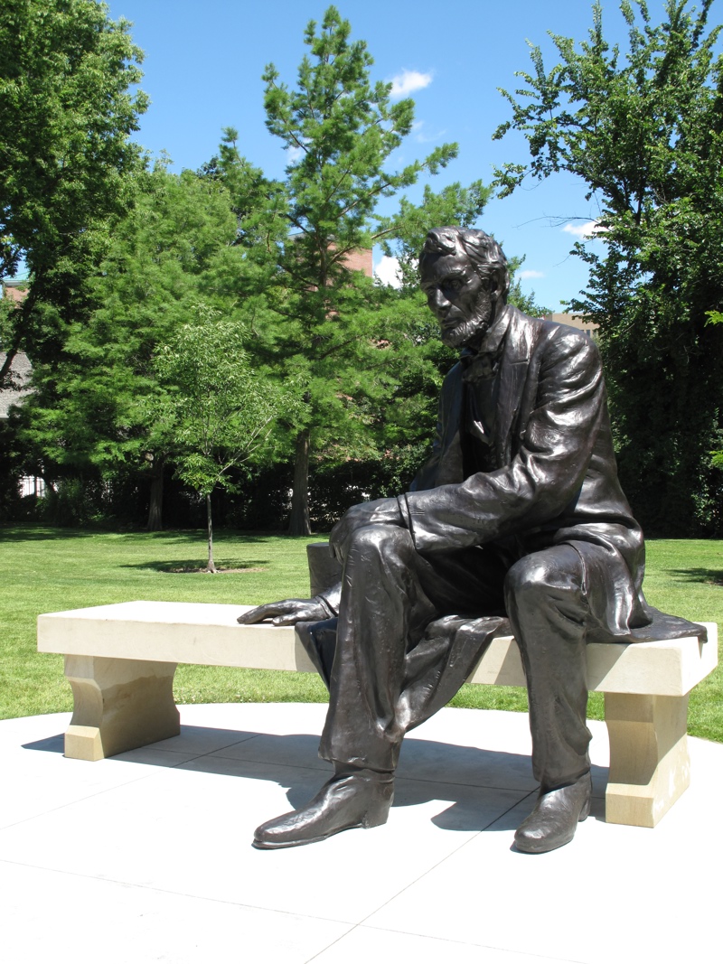 A larger bronze sculpture of President Lincoln sitting next to his signature top hat on a sandstone bench.