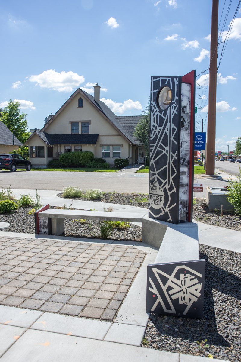 A corner bend made of cement, composite panels with printed images, and stainless steel cutouts of symbols and maps relating to Boise's first responders, namely firefighters. At the corner of the bend is a standing panel with a brass bell at its top. 