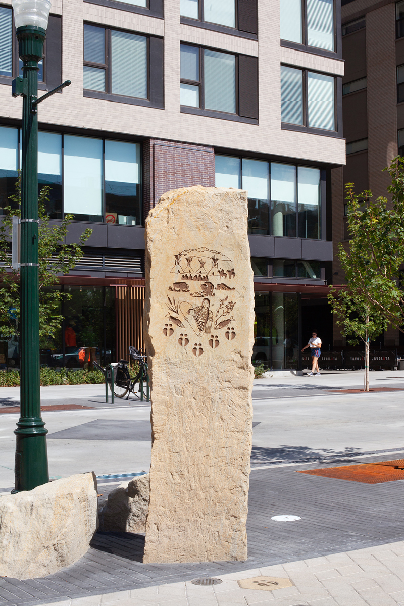 A sandstone pillar with an etching of a Native American woman of the Shoshone Bannock tribes, holding an infant in a cradle. Surrounding them are various symbols including bison hoofprints, fish, maize, teepees, figures on horseback, and mountains. 
