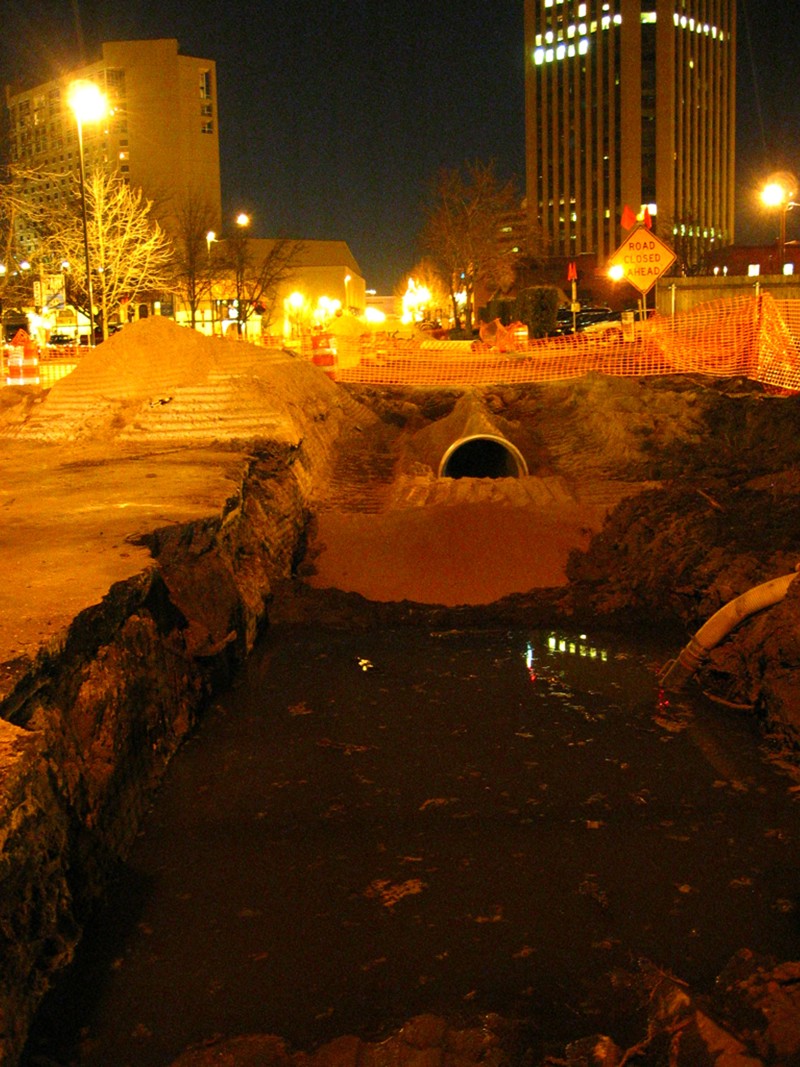 The construction site of a drainage ditch on Grove Street at night. An underground pipe is exposed, surrounded by mounds of soil and orange construction fencing.