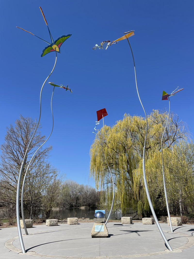 An image of the Cancer Memorial installation known as Wind Dance, located in Anne Morisson Park. The installation consists of 5 undulating steel poles, each topped with a unique and colorful kite.  At the center, between the poles, is a sandstone pedestal topped with a steel gazing globe. One the sides of the sandstone pedestal are two glass tile mosaics.