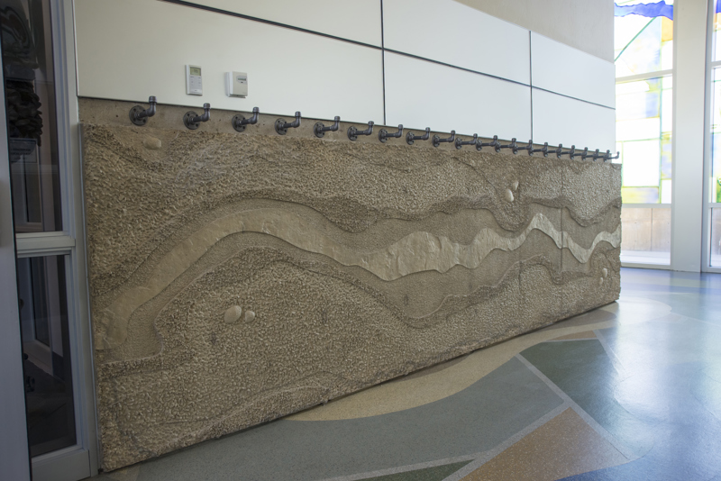 The interior side of the Boise WaterShed building. Along the bottom four feet of the building is a cement textural installation that resembles a birds-eye view or map of a river and its previous shorelines that have shifted over time. Along the top edge of the installation are twenty coat hangers made from metal water pipes.