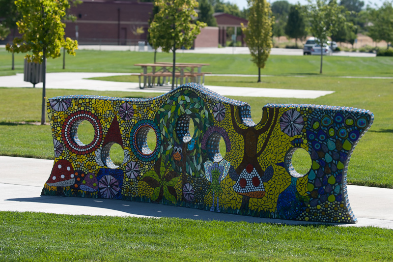 A short wall featuring a glass and tile mosaic surface. Along the wall are seven holes for peering through. The mosaic design showcases a gnome and woodland creatures with the placement of the holes aligning with the figures' heads, creating a playful "face-in-hole" effect for looking through.
