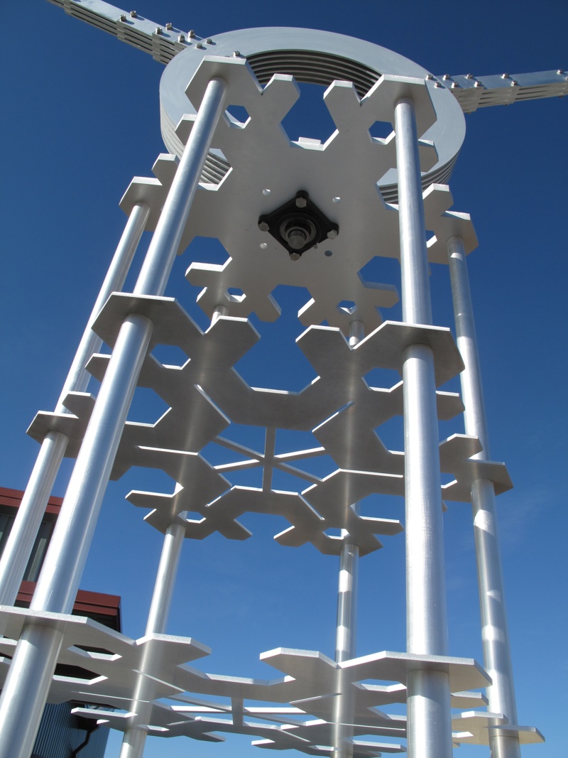 An aluminum kinetic sculpture representing a water molecule. At its central axis point is the letter O from which stems two arms with the letter H at their end. This images shows a detail closeup of the aluminum plates cut in the shape of snowflakes stacked in the support column.