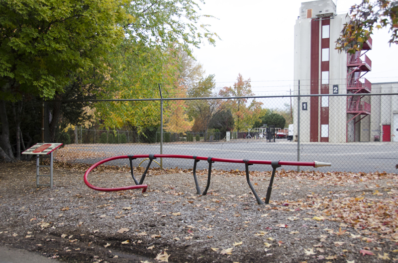A long red pipe meant for sitting on. One end of the pipe coils out of the ground, while the other end has a fire hose nozzle. The pipe is held up by steel footings with clasps that resemble hands holding a firehose. 