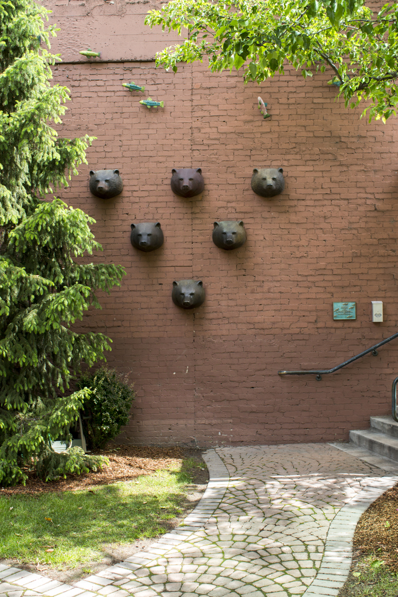 Six bronze cast bear heads mounted onto a red brick wall in a triangular patter. Above them is a school of painted ceramic fish mostly facing right to left.