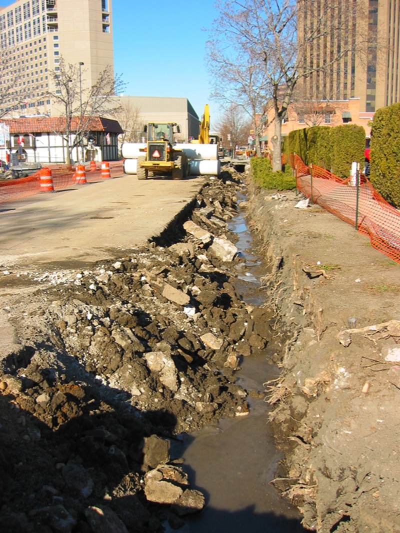 The construction site of a drainage ditch on Grove Street. An underground pipe is exposed, surrounded by mounds of soil and orange construction fencing.