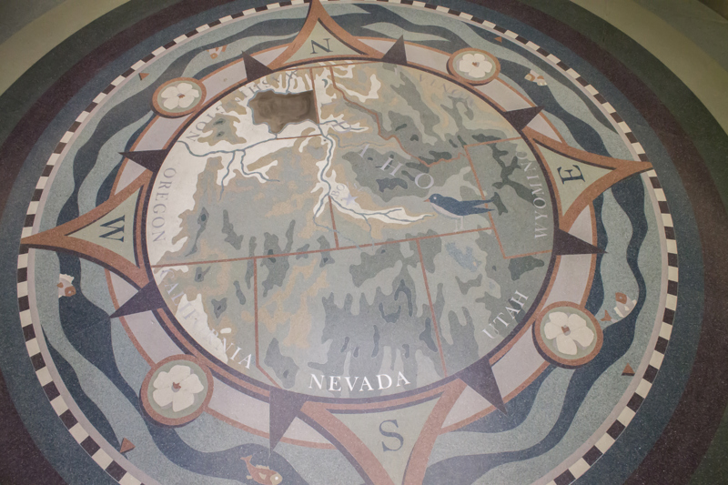 A terrazzo floor installation of a rose compass. In the middle of the compass is a map of the Northwest United States. Idaho is the only state in full view with Boise at the compass's center. The names of all the states that border Idaho (Washington, Oregon, California, Nevada, Utah, Wyoming, Montana) are written along the inner ring of the compass. On the outside of the compass, in the location of the intermediate directions, are four syringa flowers. Encircling the entire compass are fish in a